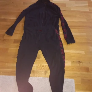 Svart hoodie från Hugo med röd logga - Svart hoodie från Hugo med röd logga längs ärmarna och dragkedja framtill. Tröjan har långärmad design och ribbade muddar. Materialet är mjuk bomull och polyester, perfekt för chill dagar. Snyggt snörningsband i midjan och cool streetstil.