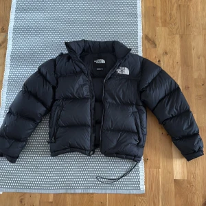 Svart dunjacka The North Face XS - Svart dunjacka från The North Face i storlek XS. Jackan är pösig och har en klassisk pufferstil med dragkedja, hög krage och innerficka med dragkedja. Perfekt för kalla dagar och har logga på både bröstet och ryggen. Materialet är polyester och dun. Två små hål som man knappast kan se på första bilden på vänster ärm och i mitten till höger. Är precis för liten för mig som är 180cm