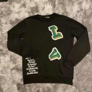 Louis vuitton sweatshirt - Svart sweatshirt från Louis vuitton med broderade bokstäver i grönt, vitt och guld på framsidan. Tröjan har rund hals, långa ärmar och vit textbrodyr vid nederkanten. Perfekt för dig som gillar streetstyle och coola detaljer.