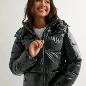 Svart glansig pufferjacka med huva - Säljer en svart glansig pufferjacka med huva och dragkedja framtill. Jackan har quiltad design, lång ärm och en riktigt snygg streetstil. Perfekt för kalla dagar när du vill hålla värmen och ändå se cool ut.