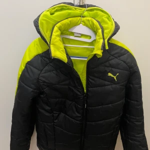 Svart och neon gul pufferjacka Puma - Säljer en fet pufferjacka från Puma i svart med neon gula detaljer och huva. Jackan har quiltad design, dragkedja framtill och Puma-logga på bröstet. Perfekt för kalla dagar när du vill sticka ut med färg och stil.