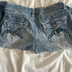 MAP lågmidjade jeans med broderade fickor - Snygga lågmidjade jeans från MAP med ljusblå tvätt och markerade vita sömmar. Bakfickorna har coolt broderade vågmönster och dekorativa knappar. Jeansen har rak passform och klassisk femficksdesign, perfekta för en avslappnad streetstil.