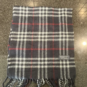 Mörkblå Rutig halsduk från Burberrys i ull - En mörkblå halsduk med vita och röda linjer. Denna vara köpte jag för två år sedan när en hemsidan skulle stänga ned så allting var på rabatt. Så jag har några burberry halsdukar till salu som du kan köpa och sälja igen sen och gå plus! Riktigt bra! Snälla köp vill bli av med den. Den är riktigt så kom inte och fråga. 