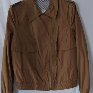 Brun bomberjacka från Mango, stl L - Snygg brun bomberjacka från Mango med dragkedja, stora fickor framtill och ribbade muddar. Jackan har klassisk krage, axelklaffar och är tillverkad i ett lätt polyestertyg. Perfekt för dig som gillar stilren och enkel design.