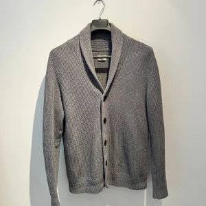 Grå kofta från Selected Homme - Snygg grå kofta från Selected Homme med ribbad struktur och bred krage. Koftan har knappar framtill och är långärmad, perfekt för lager på lager. Materialet är stickad bomull som känns mjukt och skönt mot huden. Hör av er vid frågor 