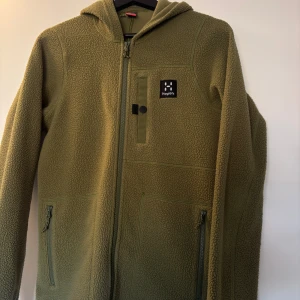 Olivgrön fleecejacka från Haglöfs XS - Mysig olivgrön fleecejacka från Haglöfs med huva och hel dragkedja. Jackan har två sidofickor med dragkedja och en bröstficka med snygg detalj och logga. Perfekt för kyliga dagar och outdoor vibes.