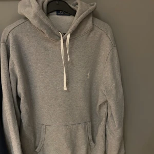 Grå hoodie från Polo Ralph Lauren - Snygg grå hoodie från Polo Ralph Lauren med vit broderad logga på bröstet. Tröjan har huva med vita snören och en stor magficka. Mjuk bomullskvalitet och klassisk passform, perfekt för chill dagar. Långärmad och stilren design.