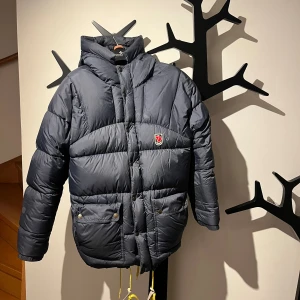 Mörkblå dunjacka från Fjällräven XXS - Säljer en mörkblå dunjacka från Fjällräven i storlek XXS. Passar även xs-m. Jackan har huva, två stora fickor med knappar och gul snörning nedtill. Klassisk Fjällräven-logga på bröstet och quiltad design som ger en schysst puffer look. En lagning på ena armen som är gjord med fjällrävens egna lagbingslappar och smälter därför in bra. Annars inga defekter.