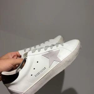 Golden Goose vita sneakers med stjärna - Snygga vita sneakers från Golden Goose med grå mockastjärna på sidan, svarta detaljer vid hälen och grå snörning. Skorna har en klassisk låg profil och platt sula, tillverkade i skinn med en stilren och urban look.