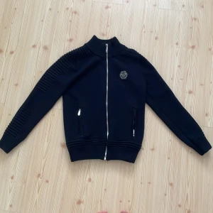 Svart Philipp Plein kofta - Svart kofta från Philipp Plein med silverfärgad dragkedja, ribbade muddar och detaljerade axlar. På ryggen finns en stor dödskalle i glittrigt mönster och framtill ett PP-logomärke. Jackan är tillverkad i bomull och polyester och har två fickor med dragkedja.Varan är äkta och använd fåtal gånger så har inte den minsta skada.Är värd Ca 13000 nypris men lägger upp den mkt billigare eftersom de plick och behöver bara pengar.Pris kan diskuteras👍