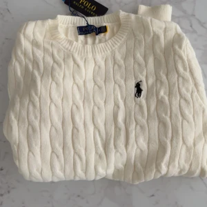 Vit kabelstickad tröja Polo Ralph Lauren - Snygg vit kabelstickad tröja från Polo Ralph Lauren med klassisk rund hals och svart broderad logga på bröstet. Tröjan har långärmad passform och är tillverkad i mjuk bomull, perfekt för dig som gillar stilrena och tidlösa plagg.