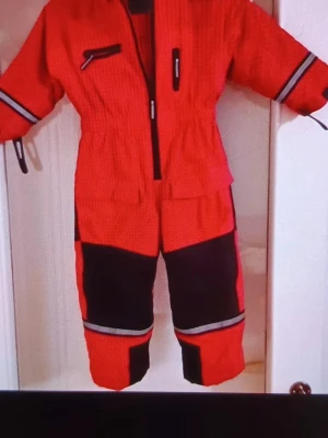 Overall barn - Säljer ett par röda täckbyxor med svarta förstärkningar på knän och ben samt reflexband. Byxorna har dragkedja framtill och är perfekta för kalla dagar. Materialet är slitstarkt och passar för vinteraktiviteter.