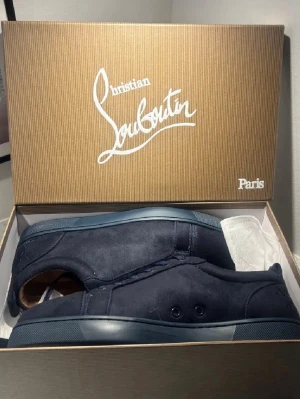 Mörkblå sneakers från Christian Louboutin - Snygga mörkblå sneakers från Christian Louboutin med klassisk röd sula och lyxig mocka på ovansidan. Skorna har rund tå, platt sula och snörning. Insidan är i beige skinn med guldfärgad logga. Perfekt för dig som vill ha något exklusivt och stilrent.