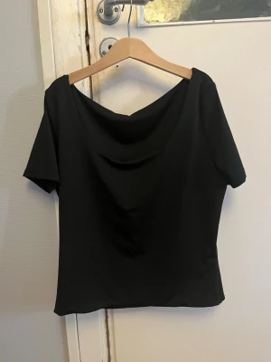 Svart topp  - En svart topp med korta ärmar och snyggt draperad urringning. Modellen är enkel men har en cool detalj framtill som ger ett extra lyft. Passar perfekt till jeans eller kjol för en clean look.