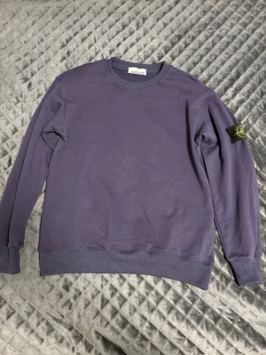 Mörklila sweatshirt Stone Island - Säljer en mörklila sweatshirt från Stone Island med klassisk rund hals och ribbade muddar. Tröjan har lång ärm och den ikoniska Stone Island-patchen på vänster ärm. Materialet är mjuk bomull, perfekt för chill dagar. Enkelt och stilrent plagg för dig som gillar streetwear.