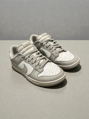 Nike Dunk Low i ljusgrå och vit - Nike Dunk Low sneakers i en clean ljusgrå och vit färgkombination. Skorna har klassisk snörning, rund tå och platt sula. Ovandelen är i skinn och textil, med Nike-logga på hälen och sidan. Perfekt för dig som gillar stilrena sneakers med retrovibe. Skorna är använda men i bra skick, de är lite smutsiga och har lite fläckar men syns inte när man har på sig dem. (Obs ej original)