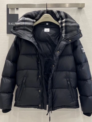 Svart pufferjacka från Burberry - Svart pufferjacka från Burberry med avtagbara ärmar, så den kan användas som väst. Jackan har en stor huva med grått och svart rutigt foder, dragkedja och knappar framtill samt två snedställda fickor med dragkedja. Perfekt för kalla dagar och riktigt snygg streetstil. Finns i båda storlek M och S.