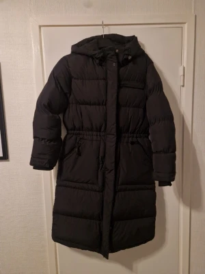 Svart lång pufferjacka med huva - Säljer en svart pufferjacka med huva och dragkedja. Jackan är lång, har quiltad design och praktiska fickor med dragkedja. Perfekt för kalla dagar och har en snygg, enkel look som funkar till många stilar.