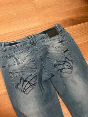 Blå bootcut jeans från Phobia - Snygga ljusblå jeans från Phobia med slitningar på benen. Bakfickorna har broderade mönster 