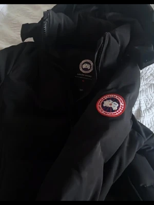 Canada Goose Vinterjacka - Svart dunjacka från Canada Goose i storlek XS. Passar även S/M. Jackan har fyllning av vitt ankdun och fjädrar, yttertyg i polyester och bomull, samt foder i nylon. Perfekt för kalla vinterdagar och har klassisk Canada Goose-logga.