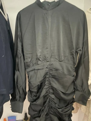 Svart omlottklänning från Eight Paris - Snygg svart långklänning från Eight Paris med hög hals och långa ärmar. Klänningen har ett omlott- och rynkat parti framtill som ger en cool och trendig look. Materialet är glansigt och känns lyxigt mot huden. Perfekt för dig som gillar statement-plagg. Passar M-S