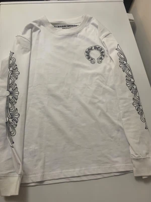 Vit långärmad Chrome Hearts tröja - Vit långärmad tröja från Chrome Hearts med svart tryck på bröstet och ryggen. Ärmarna har detaljerade svarta grafiska mönster längs sidorna. Tröjan är i bomull och har en normal passform med ribbade muddar vid ärmslut. Perfekt för dig som gillar streetwear och snygga detaljer.
