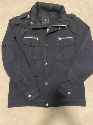 Knappt använd mörkblå fieldjacket från Riley - Snygg mörkblå fieldjacket från Riley med ståkrage, silverfärgade dragkedjor på bröstfickorna och stora knappar framtill. Jackan har långärmade ärmar, axelremmar och flera fickor med både knapp och dragkedja. Perfekt för dig som gillar stilren och funktionell design.