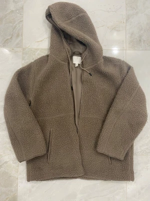 Beige teddyjacka med huva från H&M - Mysig teddyjacka i beige från H&M, storlek M. Jackan har huva med snörning, dragkedja framtill och två fickor med dragkedja. Perfekt för kyliga dagar och har en loose passform som ger en chill vibe.