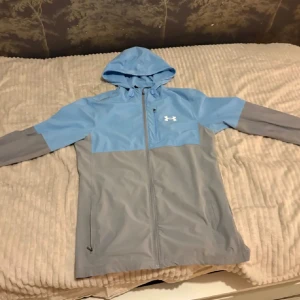Blå och grå vindjacka från Under Armour - Snygg vindjacka från Under Armour i blått och grått med huva och dragkedja hela vägen. Jackan har långärmade ärmar och två sidofickor med dragkedja. Perfekt för dig som gillar sportig stil och vill ha något lätt och funktionellt. Liten skavanker vid dragkedjan men funkar bra ändå