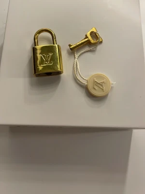  guldpläterat lås med nyckel -  guldpläterat lås med tillhörande nyckel och vit tagg med logga. Perfekt som accessoar till din väska. Obs, vet ej äktheten så prissätts som icke-org! Skick syns på bilden, men fungerar som den ska.