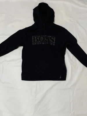 En svart Hugo Boss hudie  - Säljer en svart Hugo Boss hudie med svarta glänsande stenar på Hugo Boss namnet. Med tiden har den krympt från storlek L till M