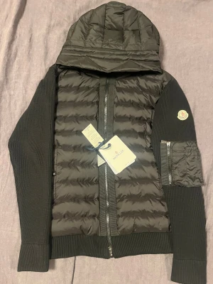 Svart Moncler pufferjacka med huva (har kvitto)  - Svart pufferjacka från Moncler med stickade ärmar och quiltad front. Jackan har huva, dragkedja framtill och en ficka med dragkedja på ena ärmen. Moncler-logga på ärmen och ribbade muddar. Perfekt för dig som gillar stilren och sportig look.