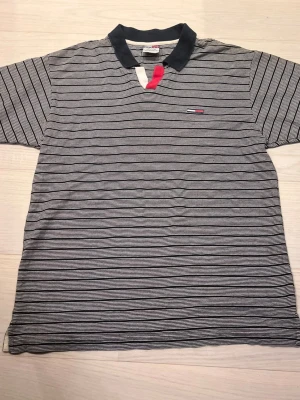 Grå Tommy Hilfiger pikétröja - Snygg grå Tommy Hilfiger pikétröja med svarta ränder i storlek L. 100% bomull. Använd fåtal gånger och skicket är mycket bra, inga slitningar eller defekter finns. Den har marinblå krage utan knappar och är riktig stilig. Skick: 8/10