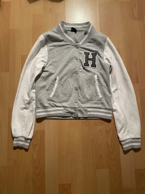 Grå och vit kofta från Gina Tricot - En collegeinspirerad kofta som är grå med vita ärmar, har ribbade muddar och krage med ränder, samt tryckknappar framtill. På bröstet finns ett stort H-tryck. Två fickor på framsidan.