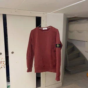 Vinröd Stone Island sweatshirt - Säljer en vinröd sweatshirt från Stone Island med klassisk patch på vänster ärm. Tröjan har rund hals, ribbade muddar och är långärmad. Perfekt för dig som gillar en klassi stil och vill ha något stilrent.