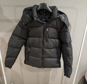 Svart pufferjacka från Polo Ralph Lauren - Svart pufferjacka från Polo Ralph Lauren med huva och dragkedja framtill. Jackan har en röd logga på bröstet, två sidofickor med dragkedja och quiltad design. Perfekt för kalla dagar och har en snygg, sportig look.