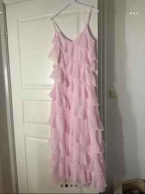 Ljusrosa volangklänning med smala axelband - Supersöt balklänning i ljusrosa med flera lager volanger och smala axelband. Klänningen har en luftig och flowy känsla, perfekt för sommarens alla tillfällen. Materialet är tunt och lätt. Djup v-ringning framtill och volanger hela vägen ner.