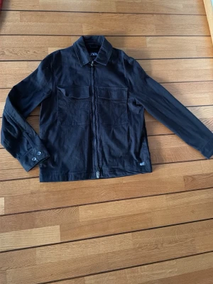 Svart overshirt från Zara med dragkedja - Svart overshirt från Zara i bomull med två stora bröstfickor och klassisk krage. Jackan har hel dragkedja framtill och långa ärmar med knapp vid mudden. Perfekt för lager-på-lager och enkel att matcha med olika outfits.