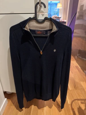 Marinblå half zip tröja från Morris - Snygg marinblå tröja från Morris med half zip och liten broderad logga på bröstet. Tröjan har lång ärm och ribbade muddar. Perfekt för dig som gillar klassisk stil med en modern touch. Materialet känns mjukt och är merino ull.