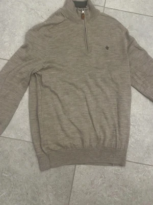 Beige stickad half zip-tröja från Morris - Snygg beige stickad tröja från Morris med half zip och hög krage. Tröjan har ett litet broderat emblem på bröstet och ribbade muddar vid ärmslut och nederkant. Perfekt för dig som gillar stilrena och klassiska plagg. Upplevs lite mindre än vanliga tröjor i S. Inga defekter!