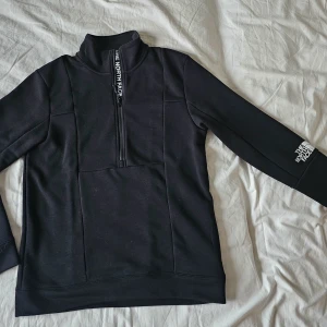 Svart half zip hoodie från The North Face - Svart hoodie från The North Face med half zip framtill och hög krage. Tröjan har lång ärm och logotypen syns både på bröstet och ärmen. Materialet är mjukt och känns som en blandning av bomull och polyester, perfekt för kyliga dagar. Helt ny! Passar till och med stl. L. Den är Oversized M. I nytt skick !