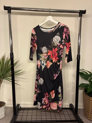Blommig svart klänning  - Svart oanvänd klänning med färgglatt blommigt mönster. Modellen har trekvartsärm och rund halsringning. Materialet är mjukt och stretchigt, vilket gör den bekväm att bära. Snygg passform som följer kroppen.
