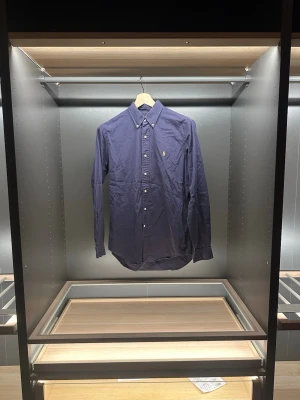 Mörkblå skjorta från Ralph Lauren - Klassisk mörkblå skjorta från Ralph Lauren i bomull. Skjortan har button-down krage, vita knappar och den ikoniska gula broderade loggan på bröstet. Långärmad och normal passform. Hör av er vid frågor eller funderingar som vanligt är priset ej hugget i sten