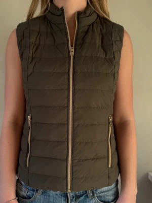 Olivgrön dunväst från Rockandblue - Snygg olivgrön dunväst från Rockandblue med quiltad design och två praktiska fickor med dragkedja. Västen har hög krage och stängs med en dragkedja framtill. Perfekt att slänga över en hoodie eller tröja för extra värme.