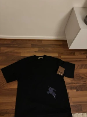 Svart t-shirt från Burberry - Säljer en svart t-shirt från Burberry med korta ärmar och ett snyggt broderat motiv i mörkblått på nedre delen. T-shirten har en klassisk passform och är tillverkad i mjuk bomull. Perfekt för dig som gillar stilrena designer med en exklusiv touch.