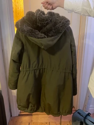 Olivgrön parkajacka Tommy Hilfiger XS - Parkajacka från Tommy Hilfiger i olivgrönt med fluffig grå fuskpäls runt huvan och quiltad gul insida. Jackan har dragsko i midjan, praktiska fickor och dragkedja framtill. Perfekt för kalla dagar och riktigt snygg stil.