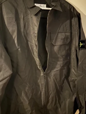 Svart overshirt från Stone Island - Svart overshirt från Stone Island med klassisk patch på ärmen. Jackan har en bröstficka med lock, krage och hel dragkedja framtill. Materialet är polyester och passformen är normal. Perfekt för dig som vill ha en clean och stilren look.