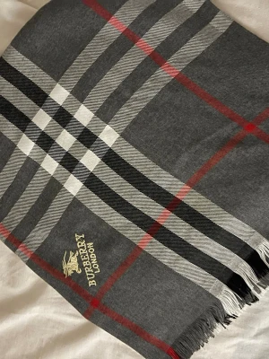 Rutig Burberry London väst i ull - Säljer en klassisk rutig väst från Burberry London med broderad logga. Västen har en snygg kombination av grått, svart, vitt och röda linjer, samt fransar längs nederkanten. Materialet är mjuk ull och designen är tidlös med den ikoniska Burberry-rutan.