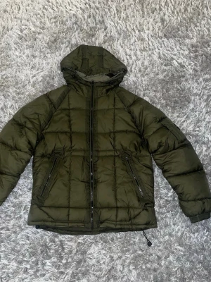 Olivgrön pufferjacka från C.P. Company - Säljer en olivgrön pufferjacka från C.P. Company med huva och två fickor med dragkedja. Jackan har ett quiltat rutmönster och en ikonisk linsdetalj på ärmen. Perfekt för kalla dagar och har en loose passform.