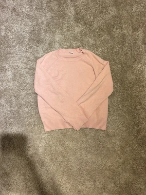 Ljusrosa stickad tröja från Lindex - Mysig ljusrosa stickad tröja från Lindex med rund hals och ribbade muddar vid ärmslut och nederkant. Tröjan har en enkel design och är långärmad, perfekt för att hålla värmen under kyliga dagar.stor i storleken